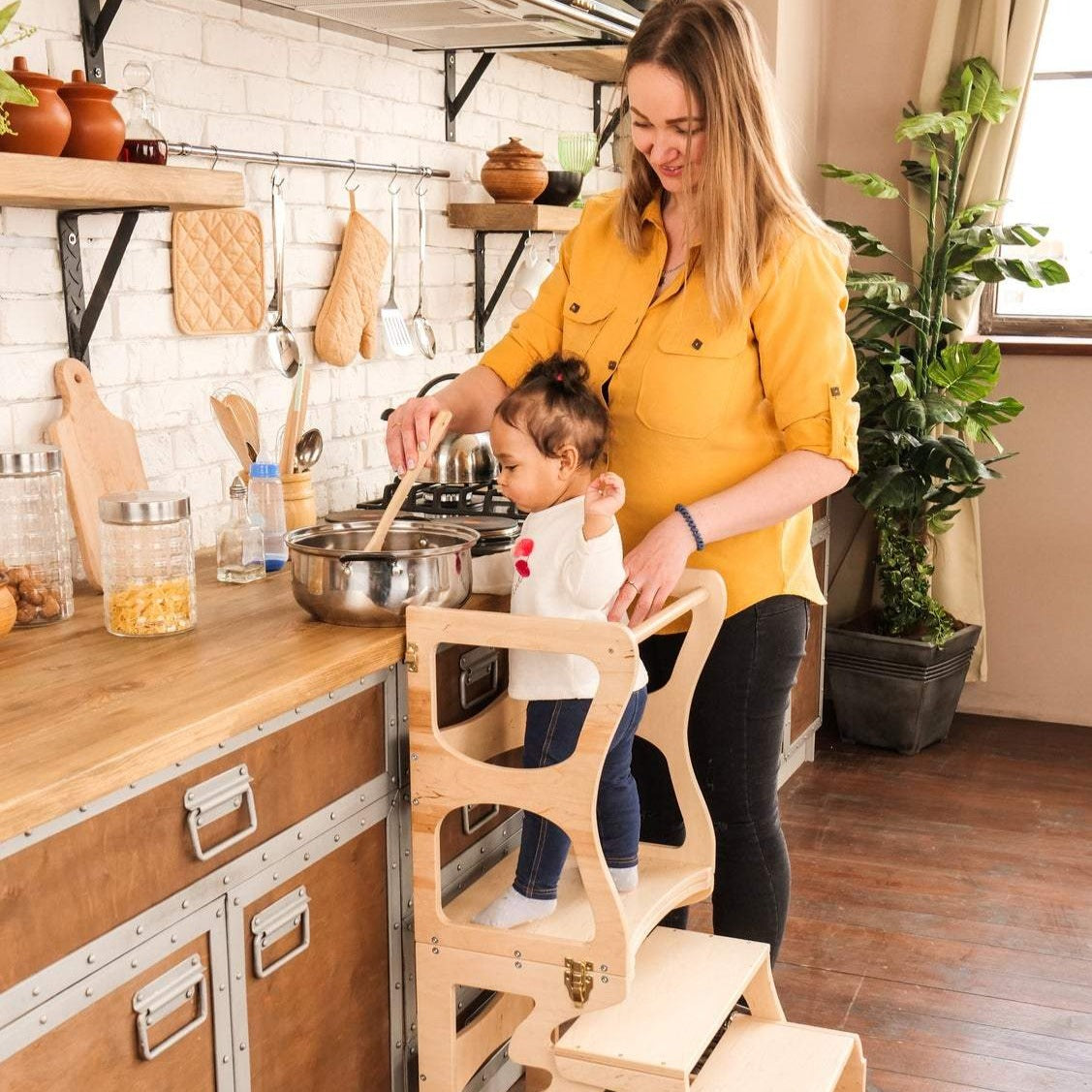 Mom with a child in the kitchen. The child stands on a high chair and reaches the countertop. Kitchen step stool chair